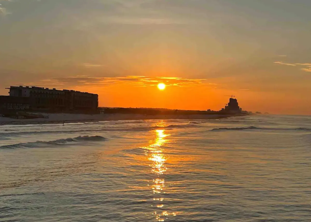 A serene view of the Gulf coastline at sunset, with gentle ocean waves reflecting warm golden and pink hues. Silhouettes of nearby buildings and the tranquil water create a peaceful, inviting scene ideal for beach walks and relaxation.