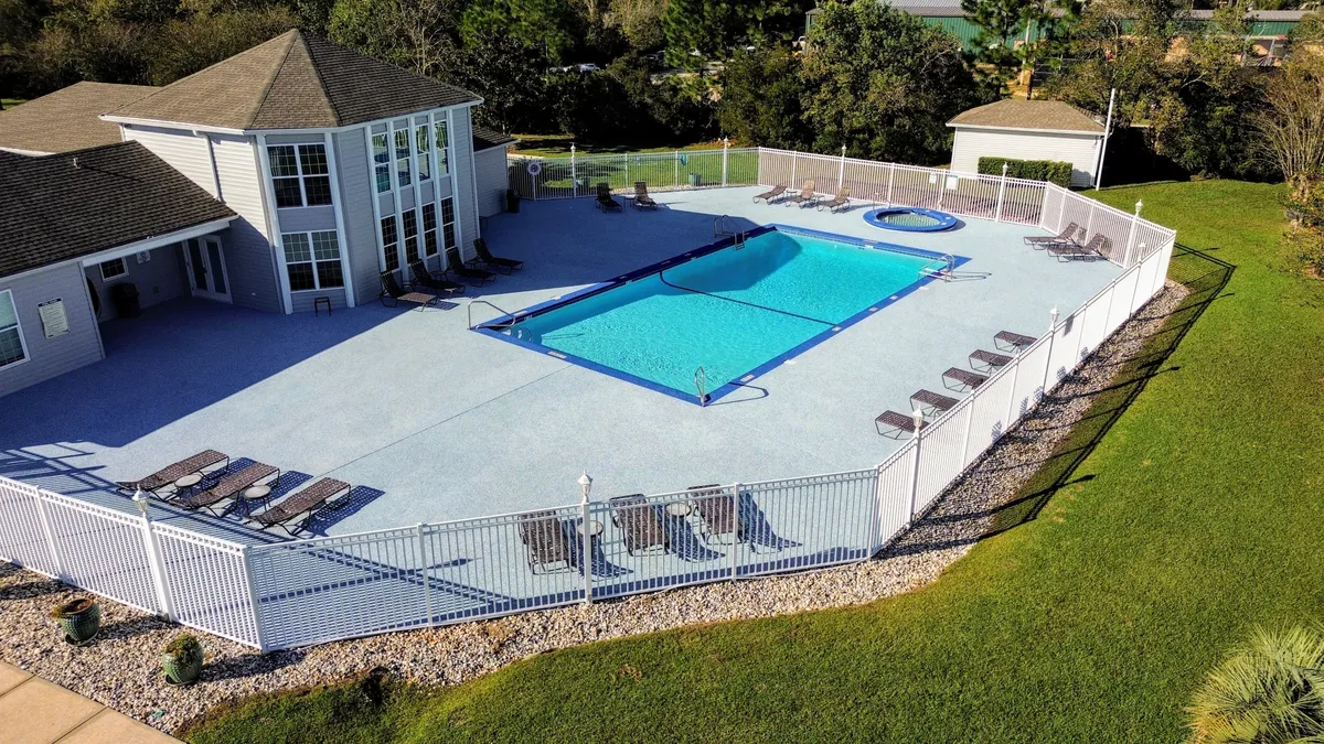 A sparkling outdoor swimming pool surrounded by lounge chairs, a secure fence, and a charming clubhouse. The scene suggests leisurely mornings, social gatherings, and a relaxing resort-style atmosphere.