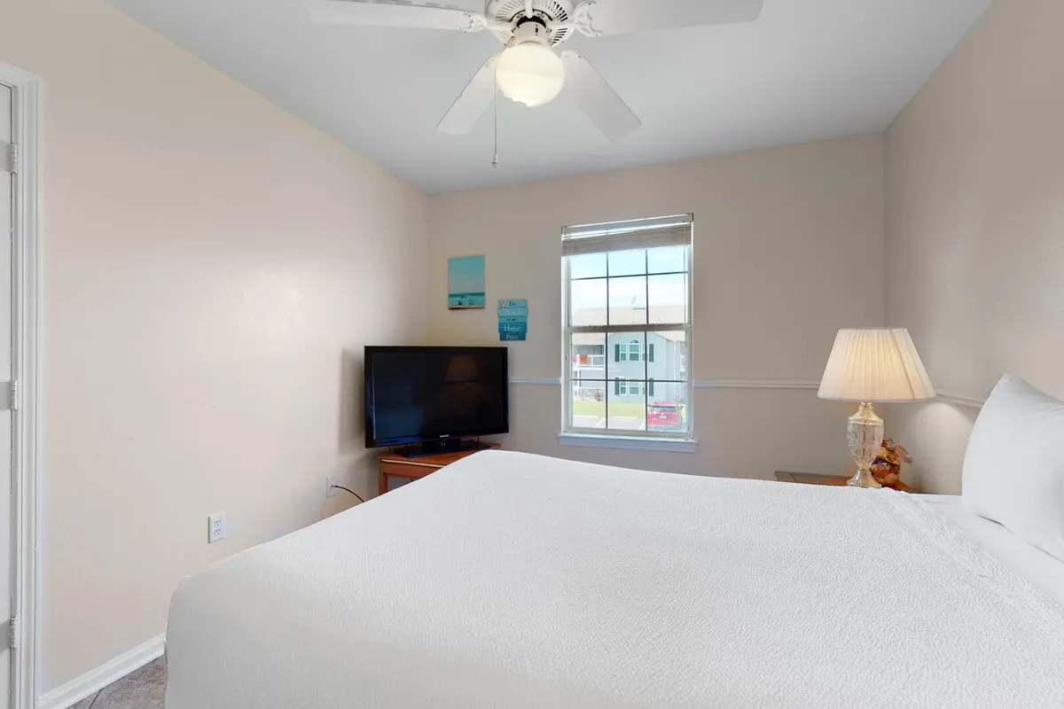 A cozy bedroom featuring a comfortable bed with soft linens, a television set on a dresser, a lamp, and a window illuminating the room with natural light. The mood is restful and inviting, perfect for winding down.