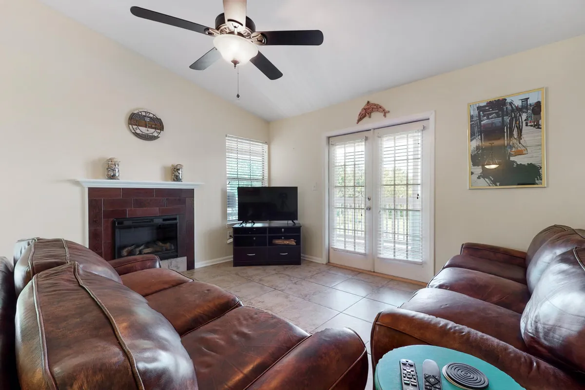 The image showcases a stylish living room with plush leather couches arranged around a cozy fireplace and a flat-screen TV, with double glass doors leading to a private balcony. The space exudes comfort and warmth, inviting guests to relax, socialize, or enjoy sunset views after a day at the beach or ballpark.