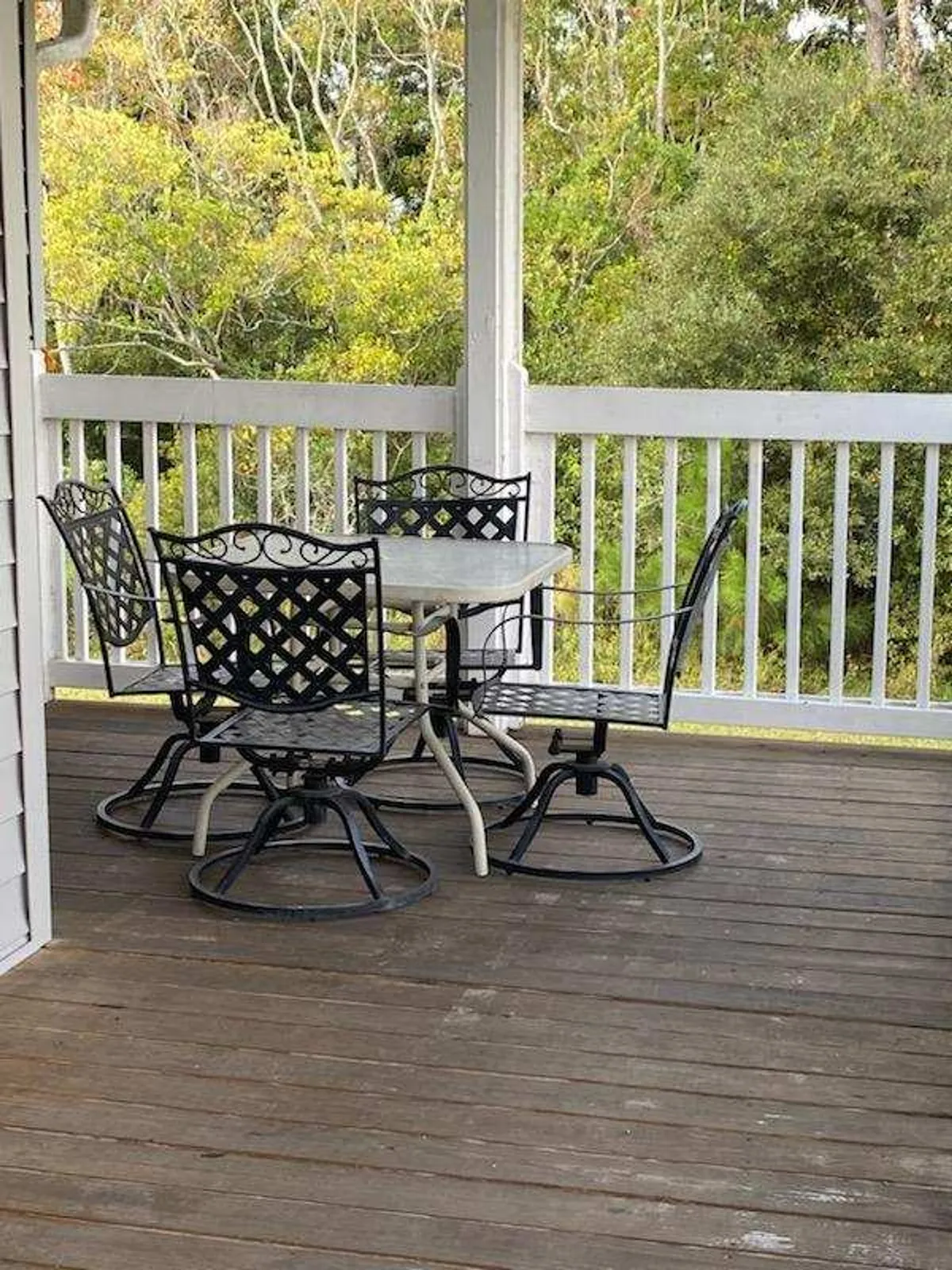 An outdoor balcony scene with black metal patio chairs and a square table, framed by a white wooden railing and lush green trees in the background. The setting is peaceful and ideal for enjoying coffee or evening refreshments.