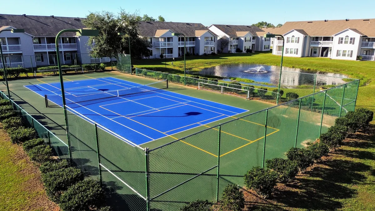 A blue tennis court with clear markings sits beside a scenic pond with a fountain, surrounded by multi-unit residential buildings and well-maintained greenery. The space is inviting for both active play and peaceful relaxation, emphasizing the blend of recreation and community.