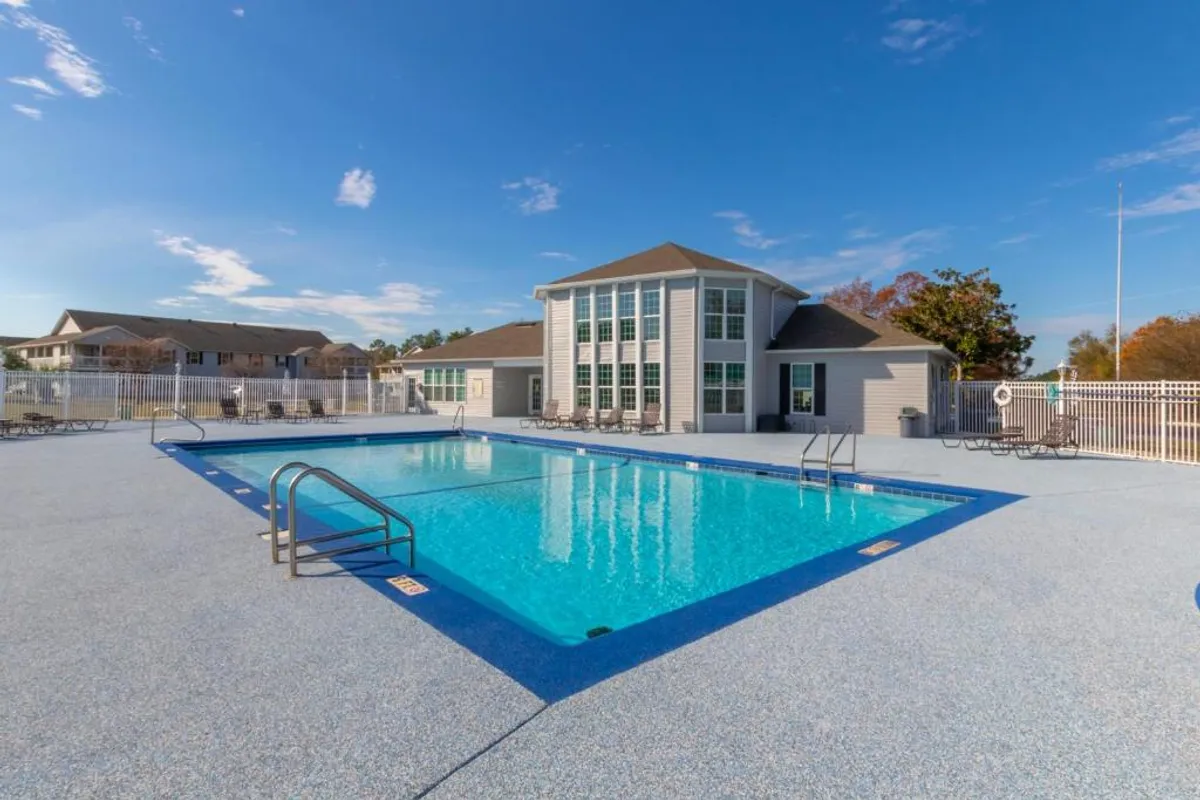 A sparkling, rectangular swimming pool surrounded by white fencing sits next to a modern clubhouse. Poolside lounge chairs are arranged for relaxation, and tall windows on the clubhouse allow for views both in and out. The setting is bright, clean, and designed for communal enjoyment.