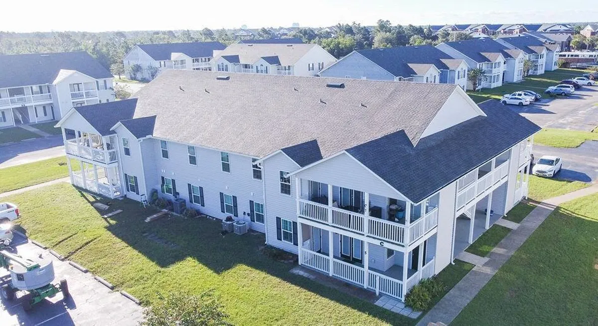 This image displays a large two-story condo building with multiple balconies, well-kept lawns, a parking area with several cars, and similar residential buildings in the background. The scene is calm and welcoming, suggesting ease of access and a friendly neighborhood environment.