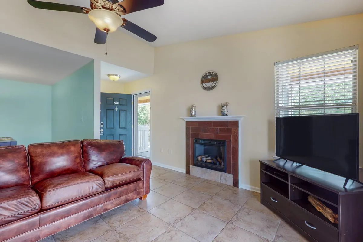 A cozy living room featuring a brown leather sofa, a decorative fireplace, and a flat-screen TV on a dark wood stand. The space is brightened by natural light streaming through the front door window, with a view and access to the balcony beyond.