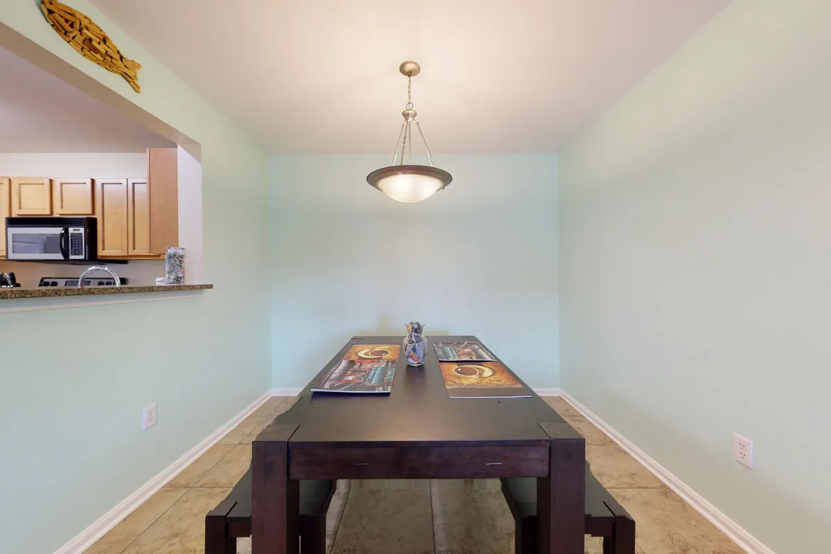 A dining area with a dark wooden table and bench seating, illuminated by a modern hanging light fixture. Light aqua walls create a fresh atmosphere, and the partial view of the adjacent kitchen (wooden cabinets, granite countertop) highlights the open-concept layout.
