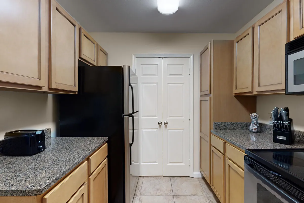 A practical and inviting kitchen interior featuring a black refrigerator, granite countertops, wooden cabinetry, and double white pantry doors. The space is clearly designed for meal preparation, cooking, and storage, creating an environment that supports group cooking and shared meals.