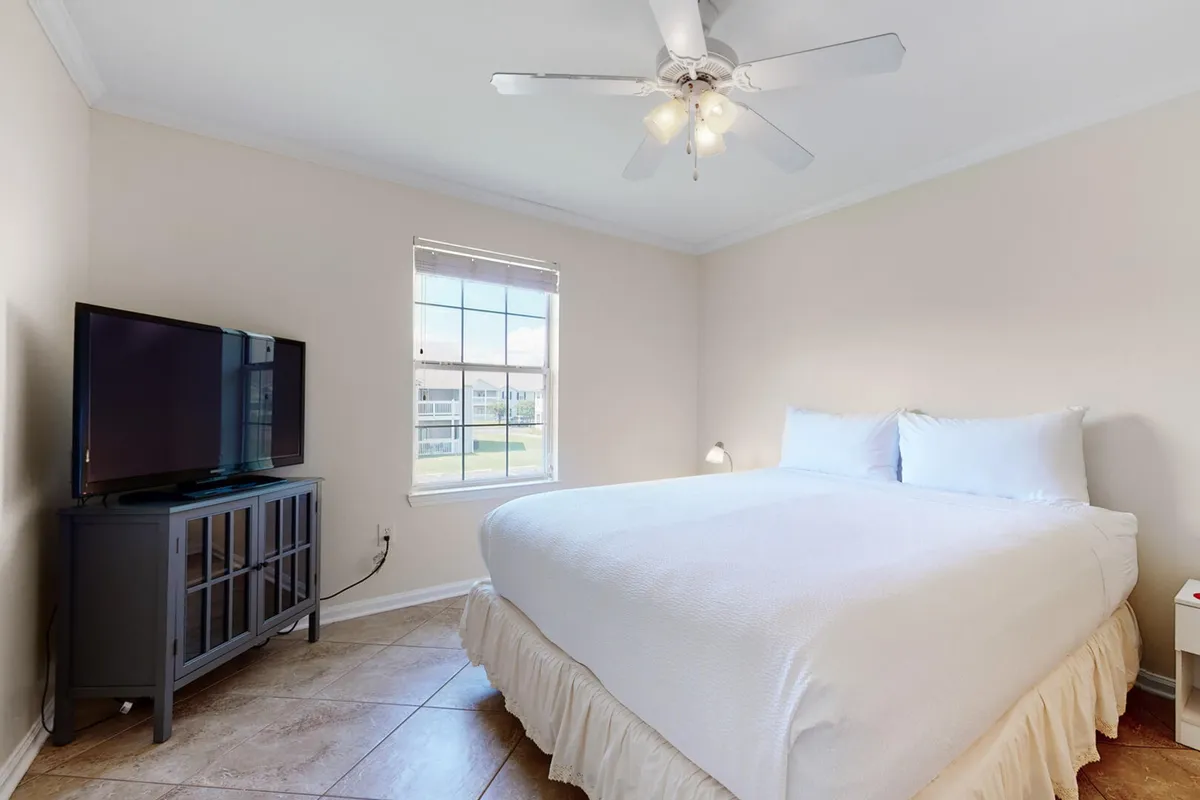 A king-size bed with crisp white linens is complemented by a flat screen TV on a dark wood cabinet, a window with natural light, and a small bedside lamp. The room conveys a sense of comfort and privacy.
