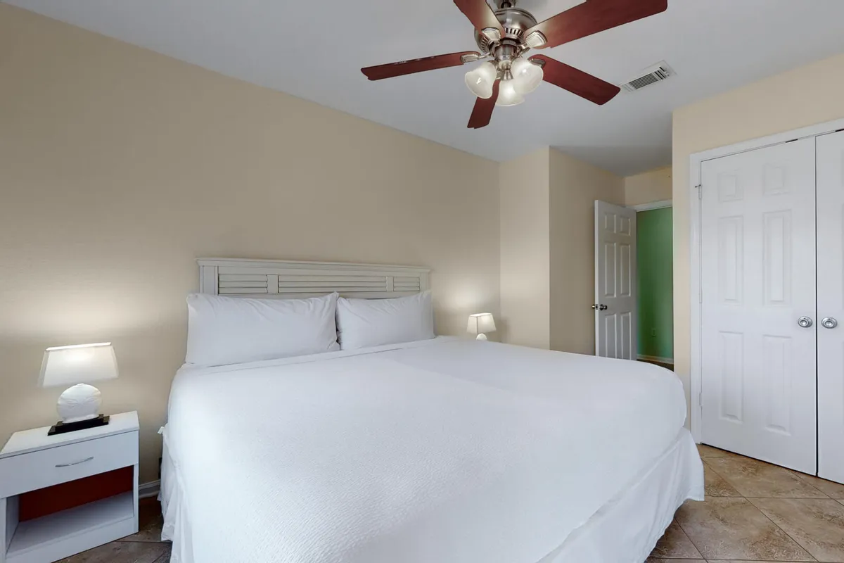 A spacious king-size bed dressed in crisp white bedding sits between matching nightstands with lamps. A ceiling fan with lights is mounted above, and a closet with double doors is visible, creating a serene and uncluttered environment.
