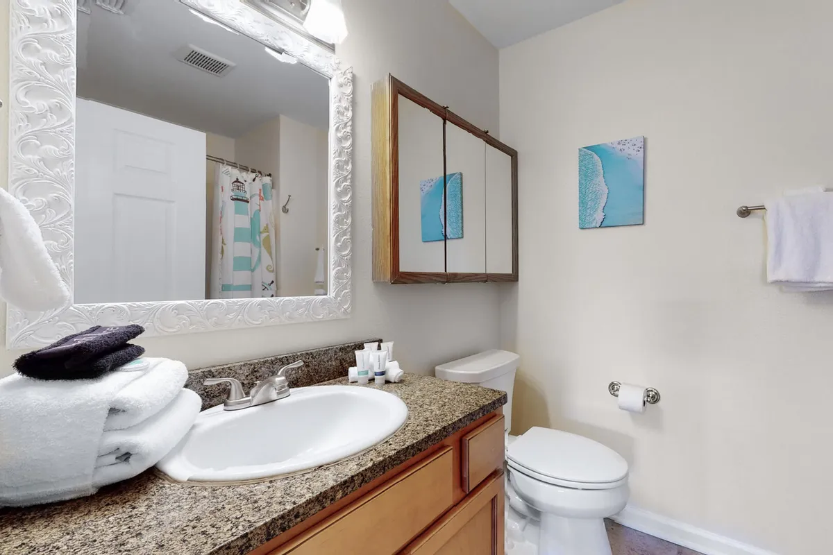 A granite-topped vanity with an ornate white-framed mirror, stacked white towels, and decorative beach-themed art creates a fresh and inviting bathroom space packed with hotel-style amenities.