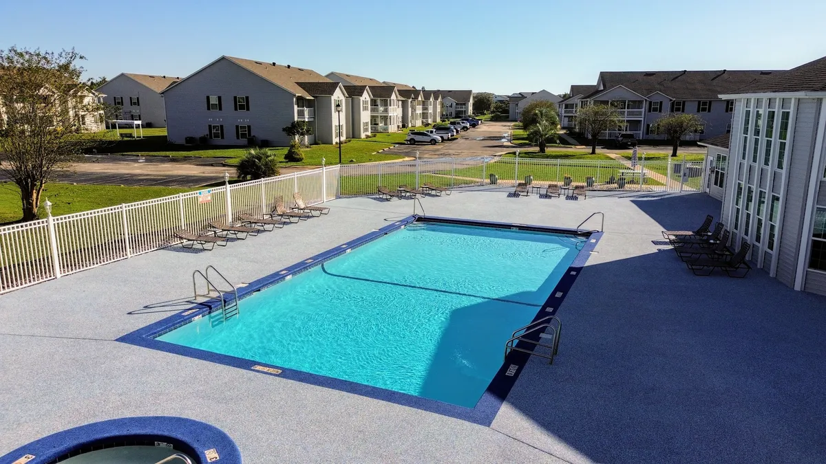 This image features an outdoor swimming pool with crystal clear blue water, a hot tub adjacent to the pool, multiple lounge chairs for sunbathing, and residential condo buildings in the background. The pool area is spacious and designed for both relaxation and socializing, with ample seating and scenic community views.