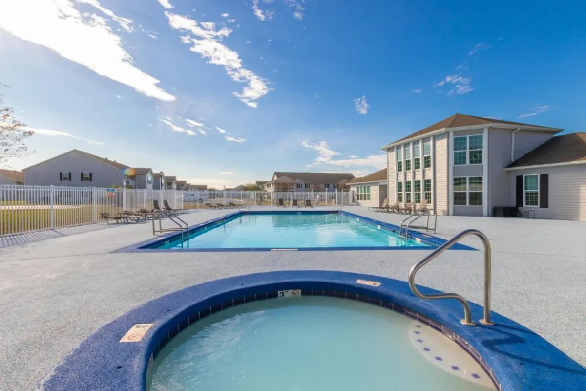 An outdoor swimming pool and hot tub are surrounded by poolside lounge chairs and a community building with large windows. The setting is bright and airy, inviting guests to relax, swim, or socialize in the clean, well-kept outdoor leisure area.