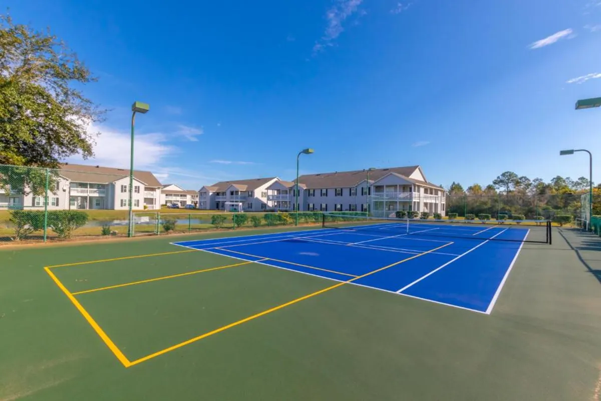 This exterior shot showcases newly resurfaced blue and green tennis courts, with clear boundary lines and tall light poles indicating evening play. Well-maintained community buildings with balconies are visible in the background, reinforcing a lively, active atmosphere.