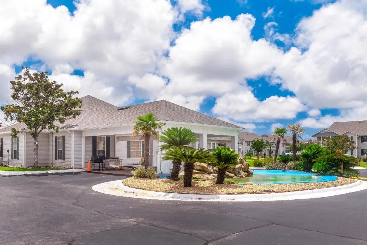 A landscaped water feature surrounded by palm and tropical plants sits in the center of a well-maintained residential community. A single-story building with white siding is visible, along with a blue sky dotted with fluffy clouds and a black asphalt road curving around the landscaping.