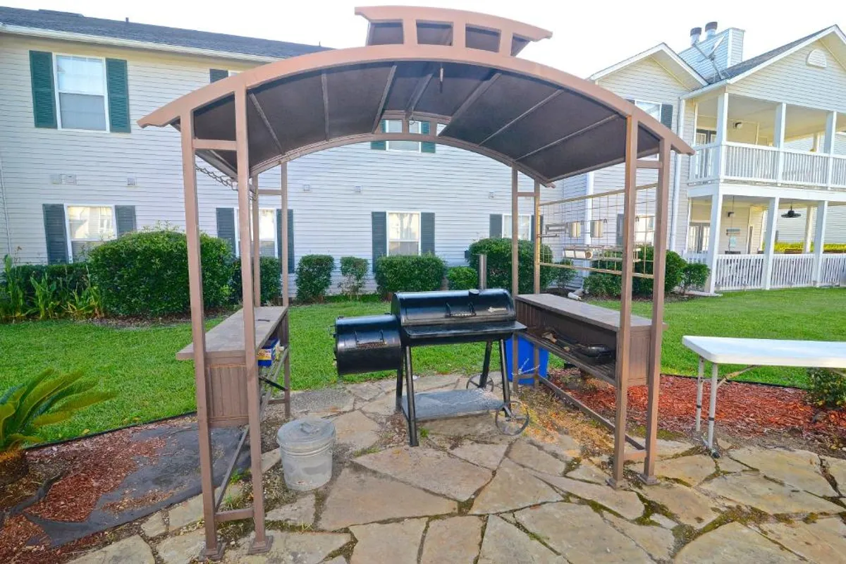 A covered barbecue grill station with a stone patio sits on a green manicured lawn, flanked by two-story residential buildings with balconies and porches. The space is clearly designed for outdoor socializing, cooking, and casual gatherings.
