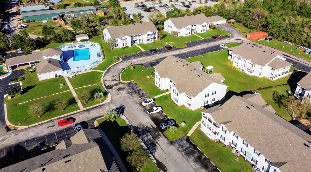 This exterior image features the community's swimming pool with an adjacent hot tub and lounge area. Several two-story residential condo buildings are visible in the background, surrounded by well-kept green lawns, walkways, and parking areas with cars. The layout conveys a sense of a vibrant, welcoming community with multiple amenities close at hand.