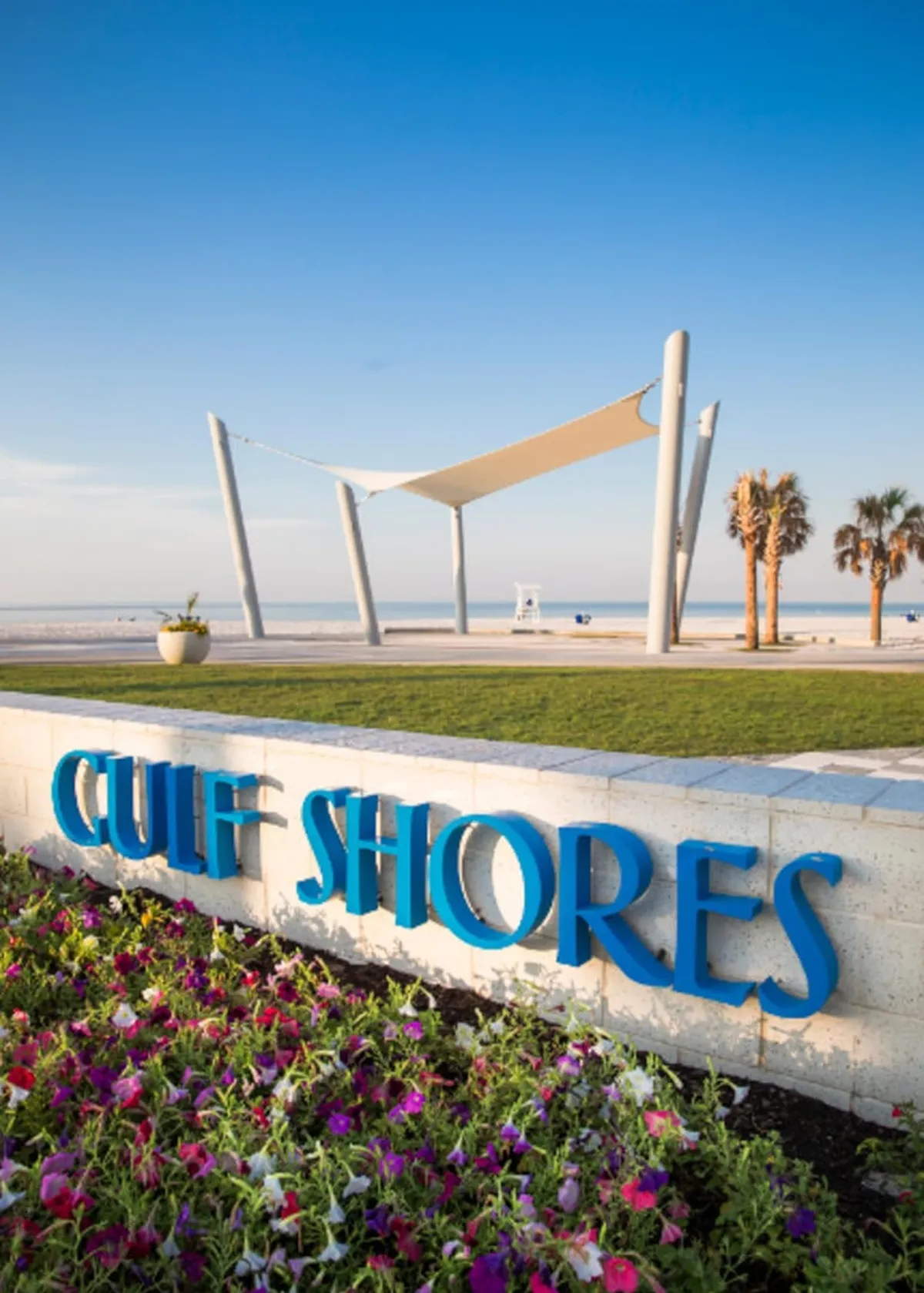 A bright blue 'Gulf Shores' sign set amid colorful flower beds and palm trees marks the entrance to the iconic beach area. A modern white shade structure stands near the sandy shoreline, with palm trees lining the background and the inviting Gulf waters just beyond the scene.